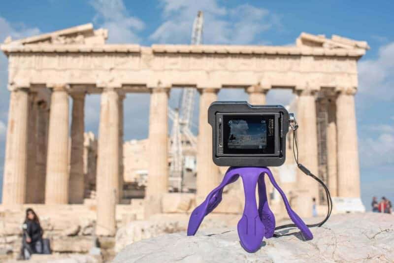 Small GoPro camera with a built-in screen mounted on a flexible purple tripod in front of the Parthenon