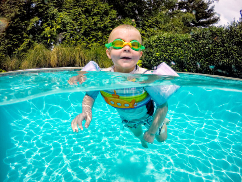 Split-level dome port shot of a goggled toddler swimming underwater in a backyard pool