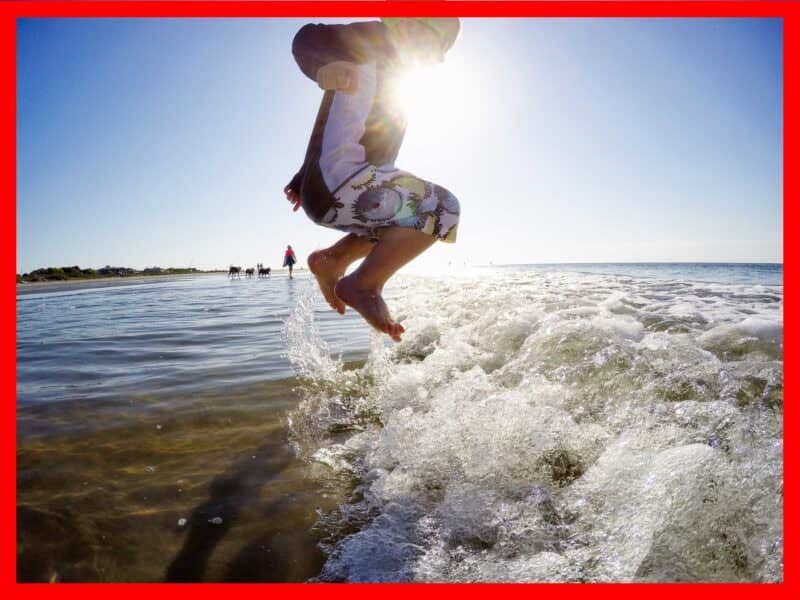 Backlit GoPro shot of a boy leaping into a breaking wave, sunlight flaring behind silhouetted legs