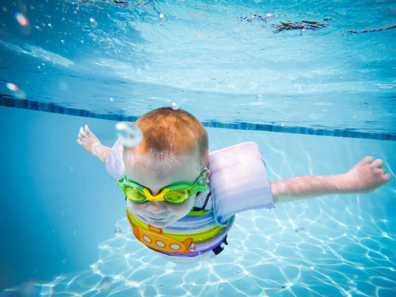 Toddler in floatie arm bands and goggles swimming fully submerged, shot with a dome port at pool level