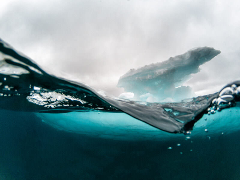 Underwater dome port split shot of an iceberg looming above the waterline in misty Arctic light