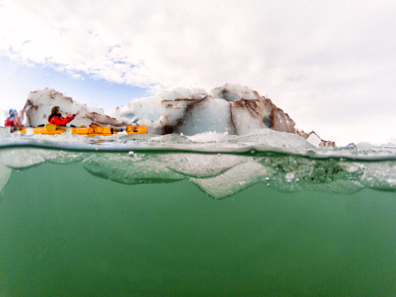 Split shot from a GoPro dome port showing kayakers paddling past a weathered iceberg in the Arctic