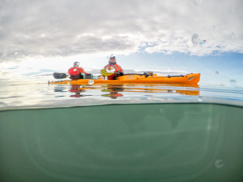 Split over/under dome shot of two kayakers on calm water beneath a cloudy sky