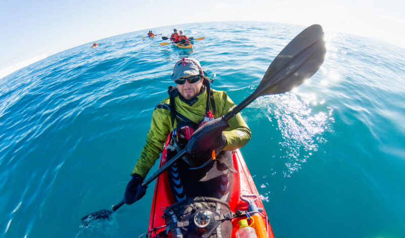 Kayaker holding up a paddle with other kayakers trailing behind on calm arctic water near Svalbard