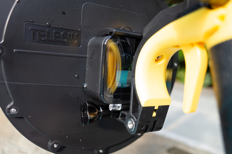 Close-up of a GoPro camera mounted inside a dome port with its clear rear door and yellow trigger