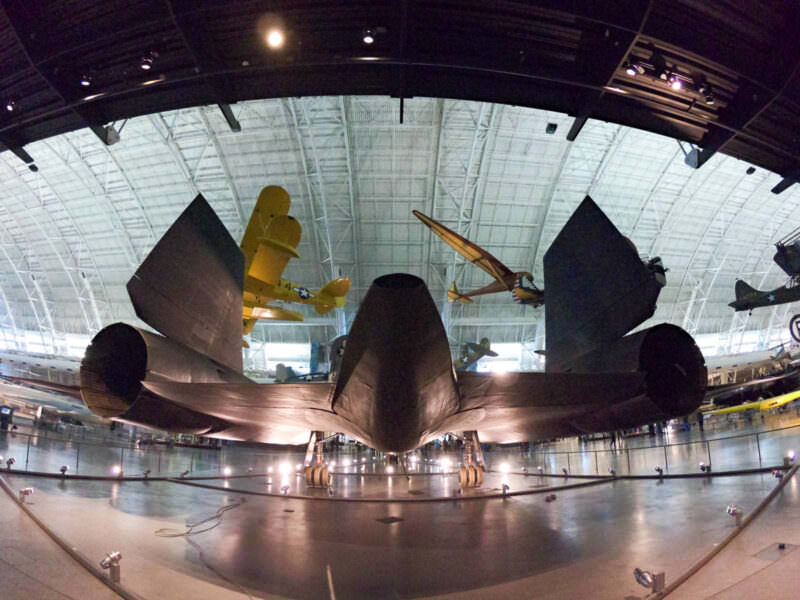 Fisheye view beneath a dark aircraft's tail fins inside a museum hangar filled with vintage planes