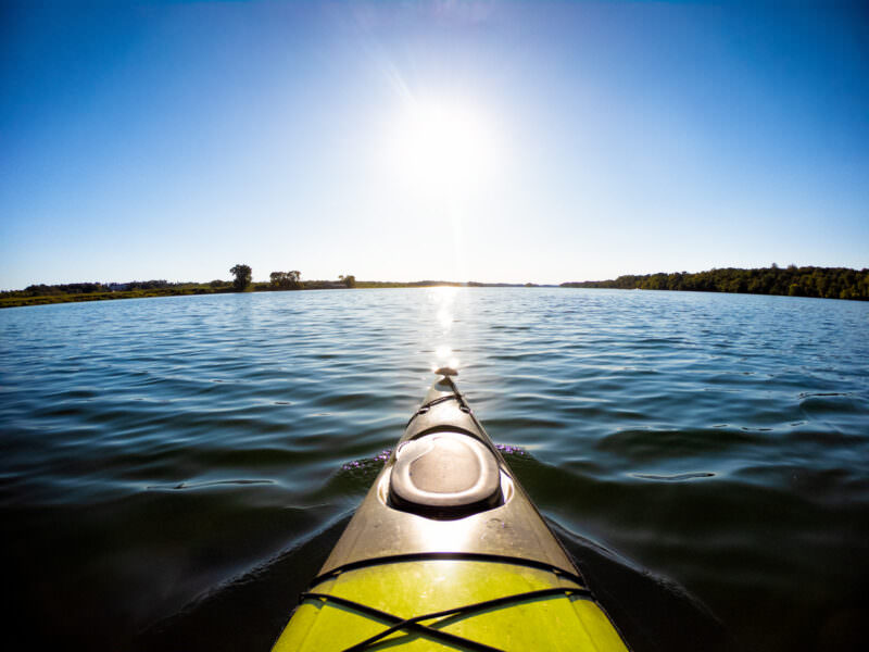 Kayak's bow pointing across a calm sunlit lake toward a tree-lined horizon