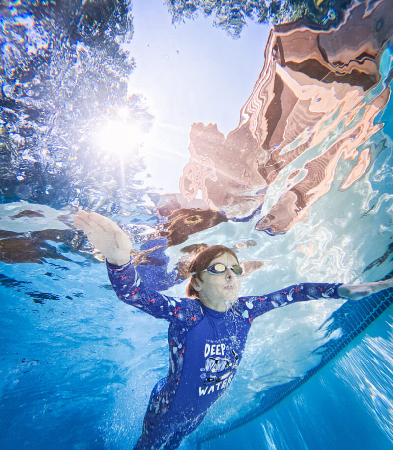 Boy in a blue rash guard swimming underwater with arms outstretched, sunlight and reflections above