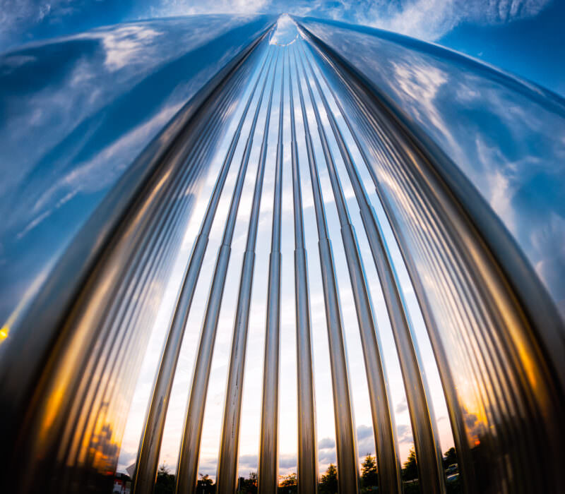 Fisheye view up through a ribbed metal sculpture toward blue sky and clouds