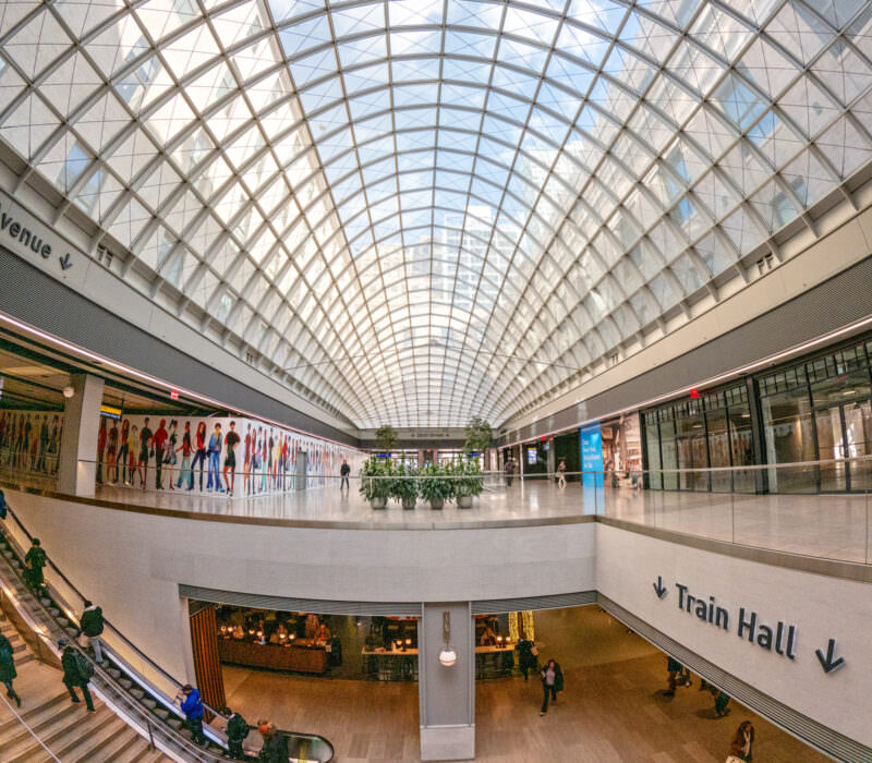 Moynihan Train Hall's glass-vaulted ceiling arching over escalators and the lower concourse in New York