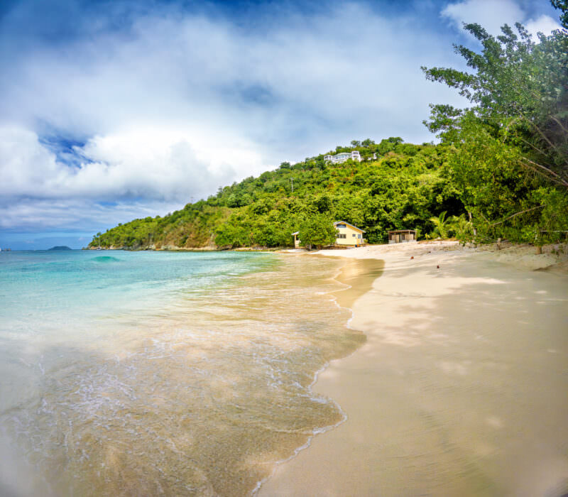Quiet crescent beach with turquoise water, pale sand, and a lone cottage beneath forested hills