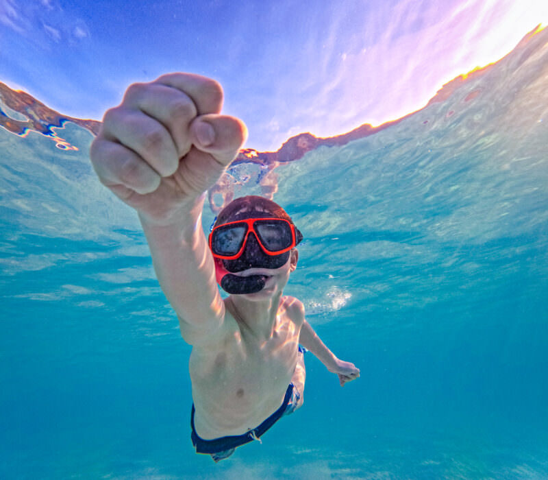Boy in red snorkel mask swimming underwater with fist raised toward the camera in clear blue sea