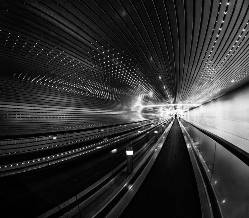 Black-and-white fisheye shot down an airport moving walkway tunnel with rows of ceiling lights