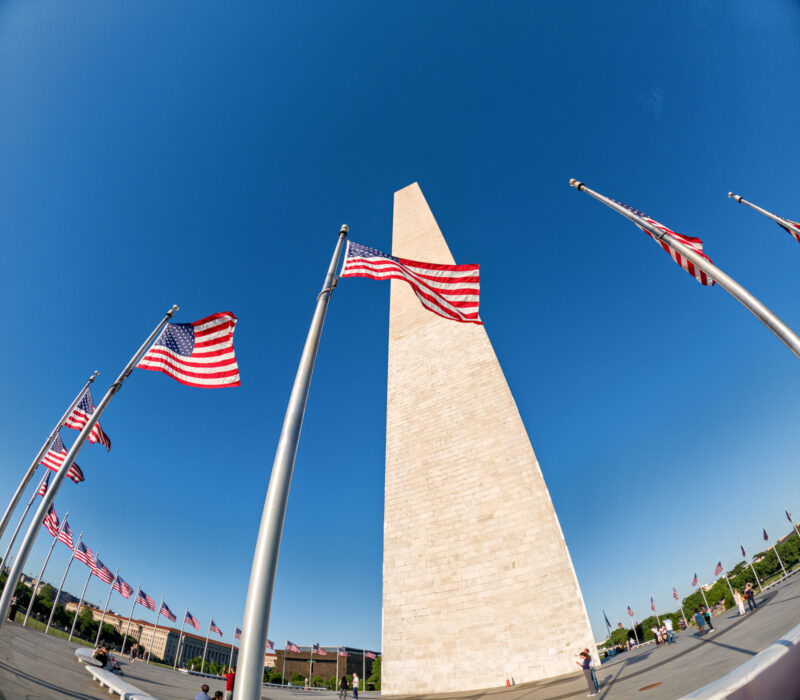 Washington Monument seen from below with American flags rippling on poles against blue sky