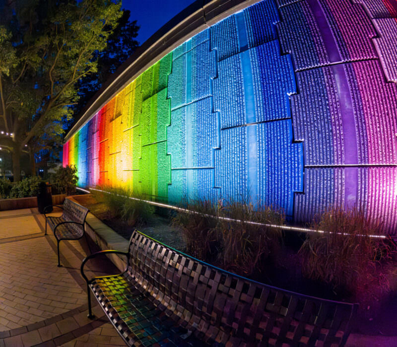 Curved rainbow-colored illuminated wall panels beside park benches at the National Landing plaza at night