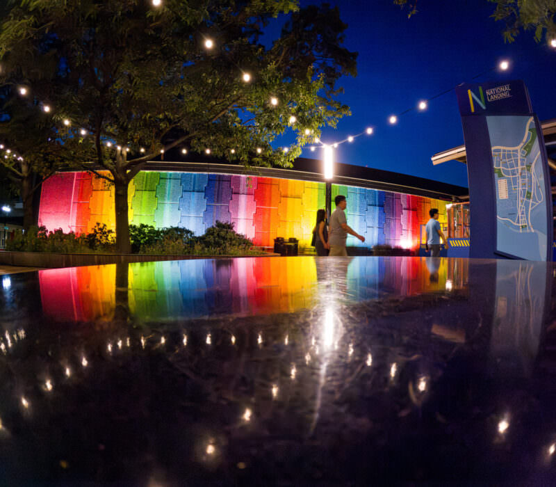 National Landing rainbow-lit wall reflected on wet pavement at night with pedestrians strolling past