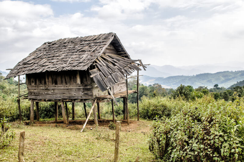 Weathered stilted rice hut with partially collapsed shingle roof, slatted wooden walls, forested hills behind