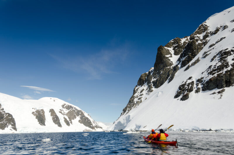 Tandem kayakers paddling along near-vertical rock and ice face at Cuverville Island, Antarctica