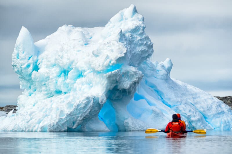 Tandem kayakers paddling beside dramatically carved blue iceberg at Petermann Island, Antarctic Peninsula