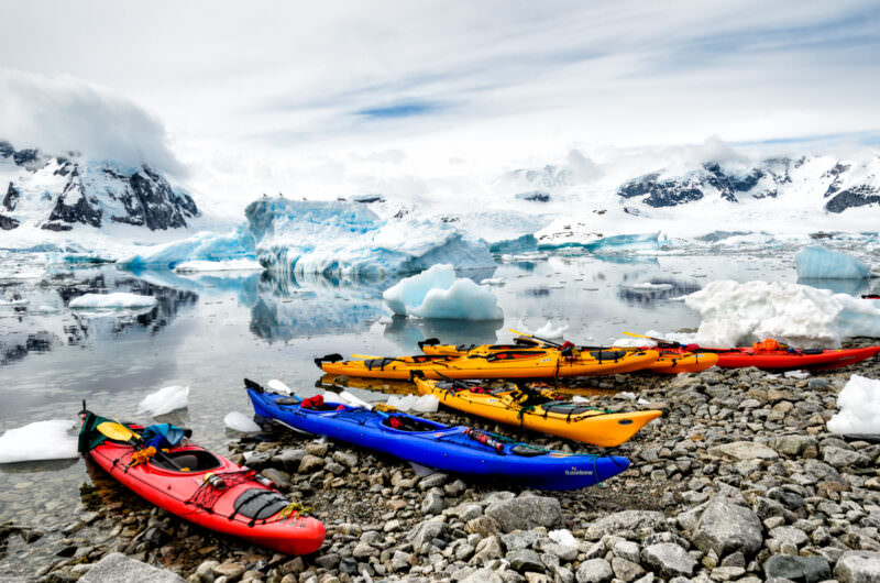 Six expedition kayaks beached on cobble shore of Cuverville Island with icebergs and mountains behind