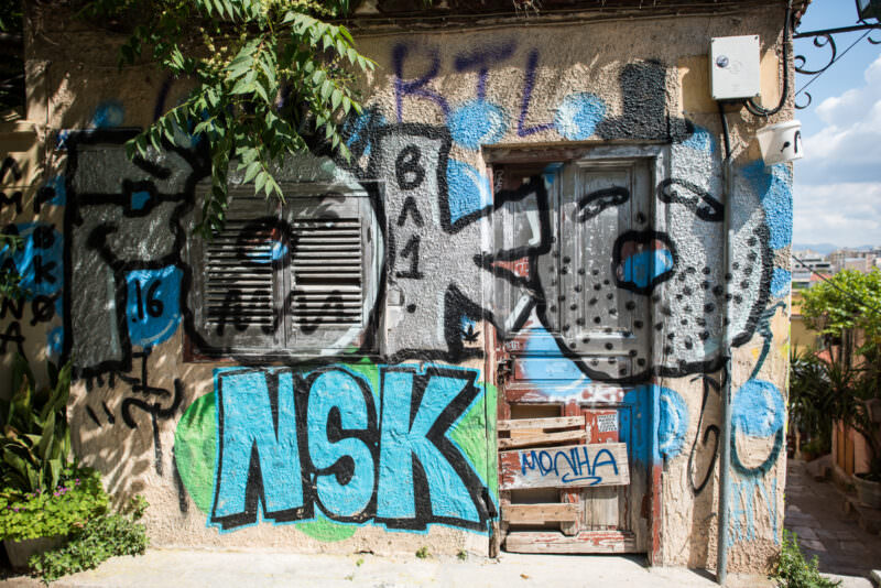Athens graffiti-covered doorway on a dilapidated building, featuring "NSK" in green bubble letters and overlapping blue and gray tags