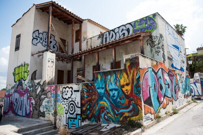 Athens graffiti covering a two-story abandoned building with colorful murals including blue flame-like faces and layered wildstyle tags