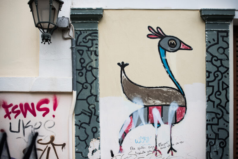 Athens street art of a minimalist stylized bird with a blue neck and red-and-black body, painted on a cream-colored wall between ornamental pilasters