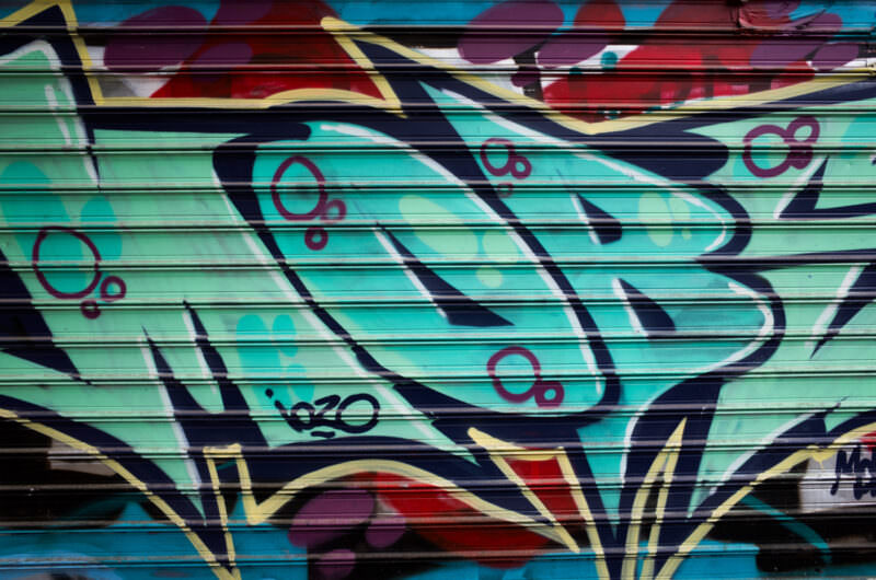 Athens graffiti detail on a metal shutter showing turquoise bubble letters with circular accents, part of a larger layered composition