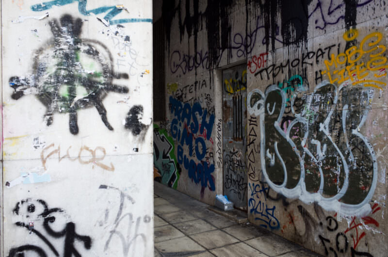 Athens graffiti layered over a concrete wall and shuttered doorway, featuring overlapping tags in black, white, turquoise, and yellow spray paint