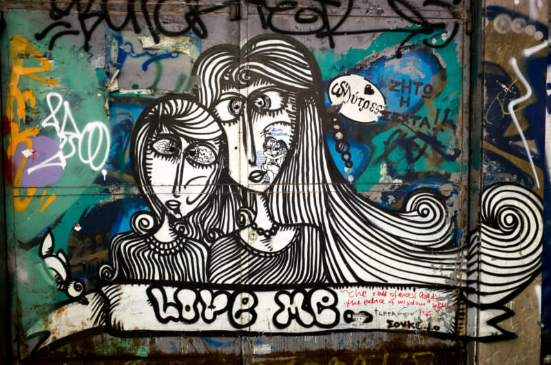 Athens street art on a wall showing two women's faces with bold black-and-white swirling patterns, signed "LOVE ΞΙΘ," amid colorful surrounding tags