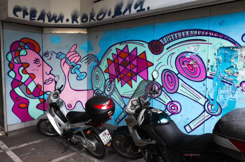 Athens street art garage mural in pink and turquoise showing a cartoon figure holding a microphone with swirling geometric patterns, two scooters parked in front