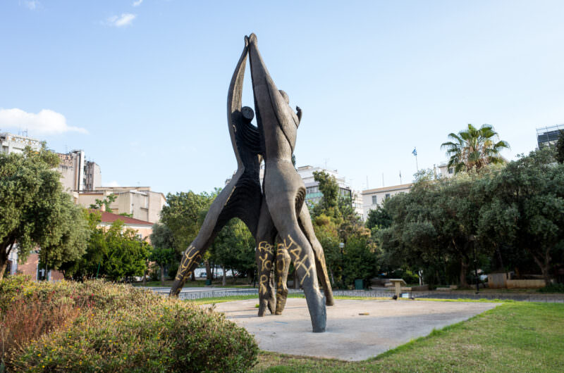 Athens public sculpture in a park of stylized bronze figures with upraised arms, silhouetted against green trees and urban buildings
