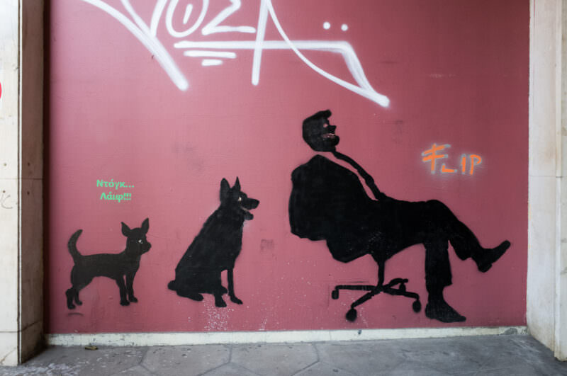 Athens stencil graffiti on a pink wall showing silhouettes of two dogs and a seated figure in black, with text in green and orange