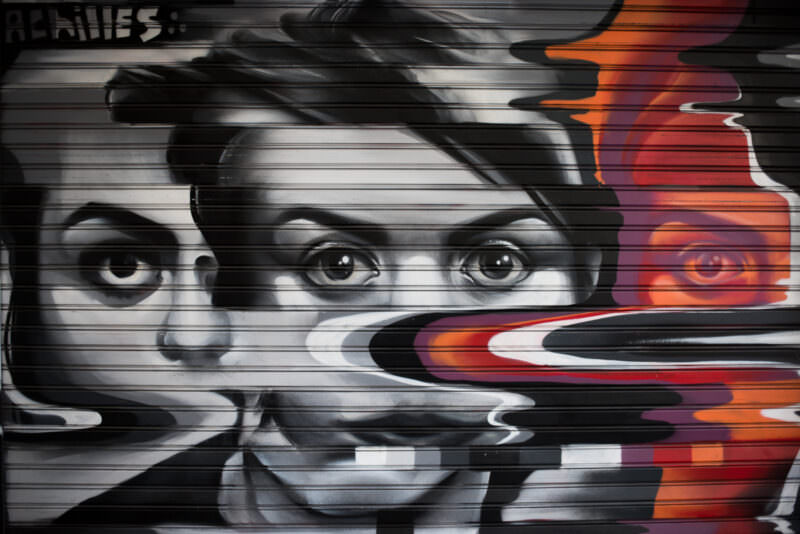 Athens street art on a shop shutter featuring surrealistic faces with melting features in grayscale, highlighted by orange-red drips