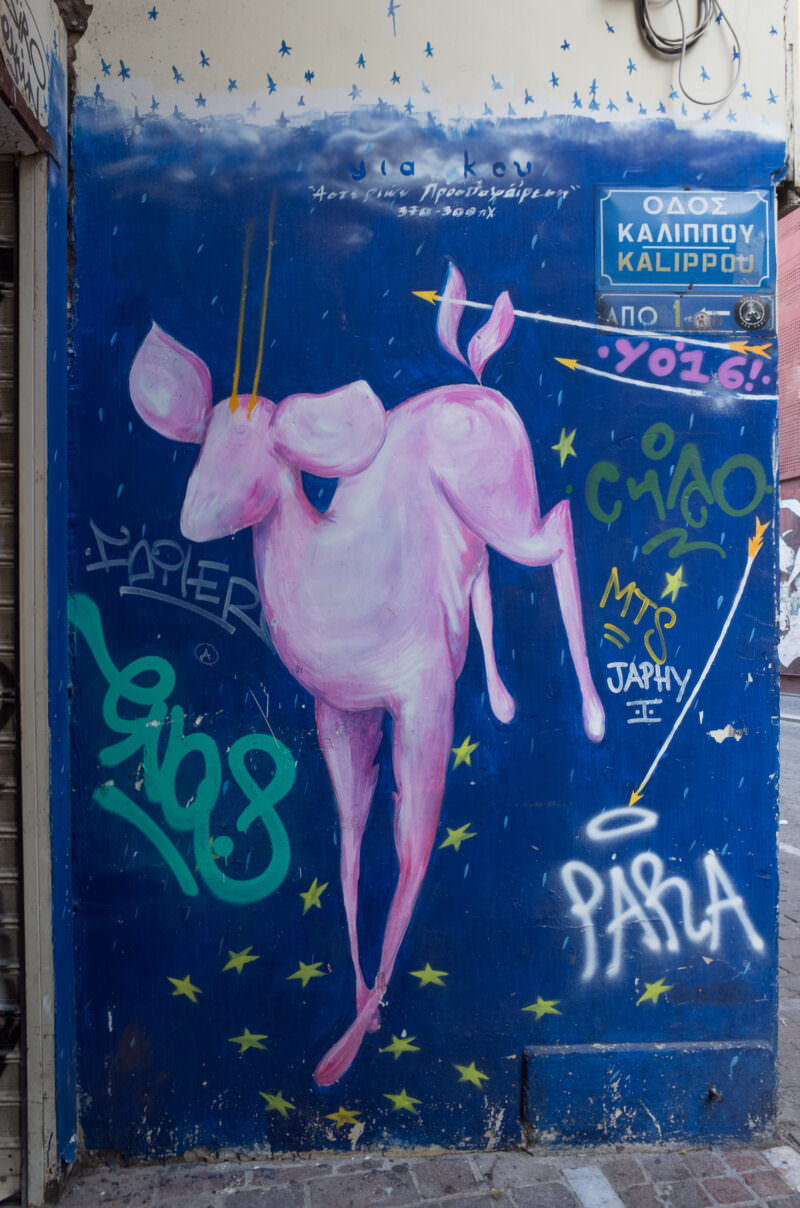 Athens mural on a blue background depicting a stylized pink winged figure pierced by arrows, surrounded by graffiti tags and Greek street signs