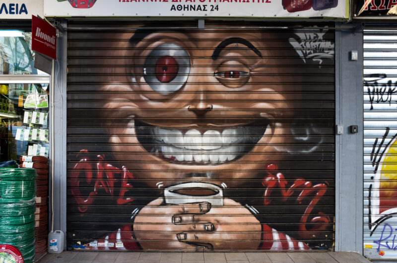 Athens mural of a grinning child in eyeglasses holding a steaming coffee cup, painted on a shop's metal shutter in brown tones