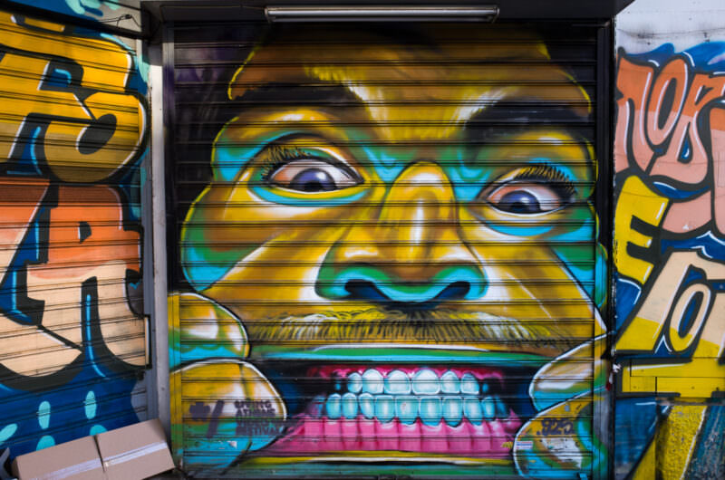 Athens street art on a shop shutter showing a wide-eyed grinning face in yellow with blue-outlined features and exaggerated teeth