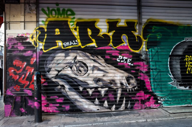 Athens graffiti mural of a stylized gray dinosaur head with sharp teeth, painted on a shop shutter beside pink and purple tags