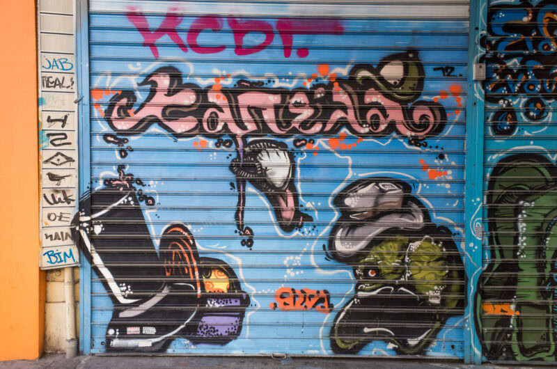 Athens mural on a blue shop shutter depicting a scooter and cursive graffiti lettering in pink, black, and orange