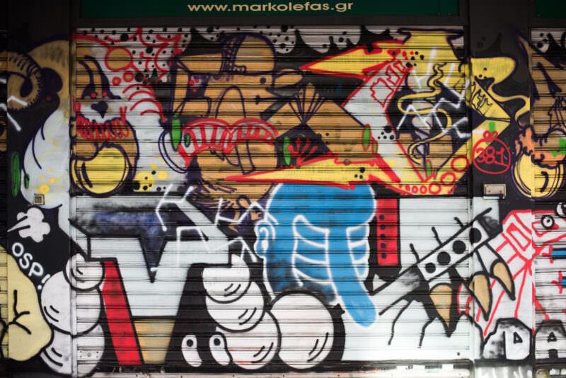 Athens street art on a shop shutter featuring stylized cartoon figures—black-and-white motorcycles and a person—amid colorful geometric patterns