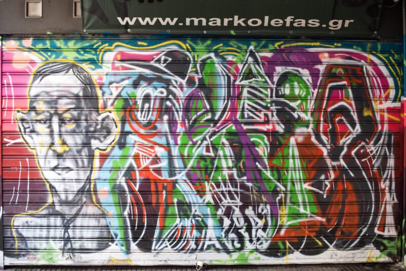 Athens street art mural on a shop shutter showing overlapping grayscale human portraits and abstract lettering in rainbow colors