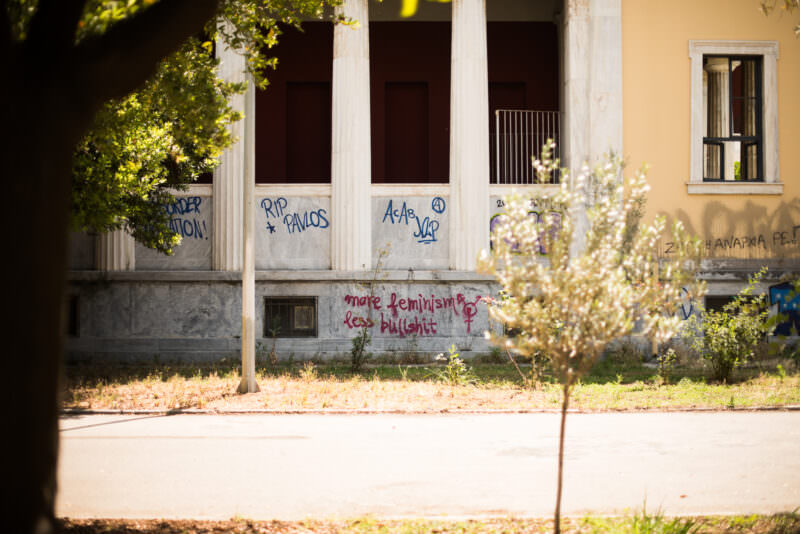 Athens graffiti on an abandoned neoclassical building facade, showing political slogans and tags painted on weathered columns in dappled sunlight