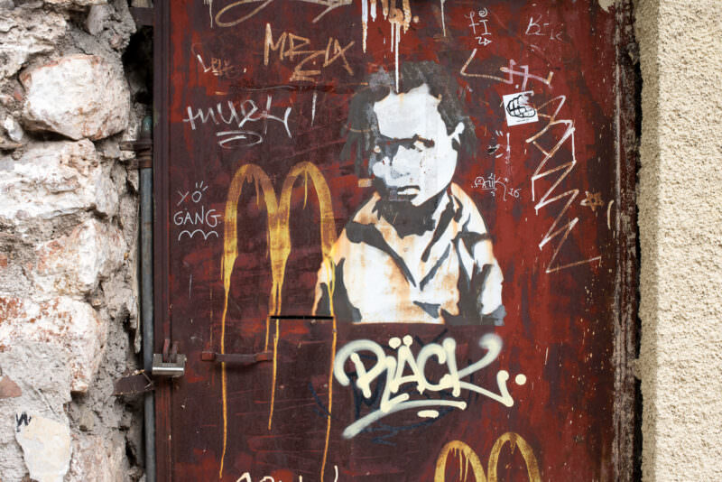Athens graffiti stencil of a figure beside golden arches and tags including "PÄCK," spray-painted on a weathered brown door