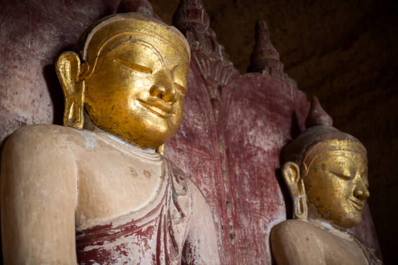 Two gilded Buddha figures in painted niche at Dhammayangyi Temple, faces densely covered in gold leaf against weathered red-painted robes