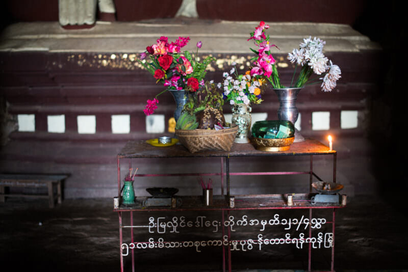 Metal offering table at Dhammayangyi Temple holding flowers, incense, candle, and fruit basket, with Burmese script on frame