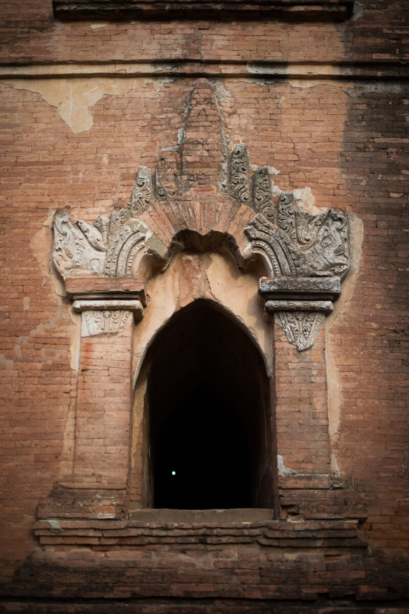 Pointed arch window at Dhammayangyi Temple framed by weathered stucco ornament with flame-like finials against red brick facade