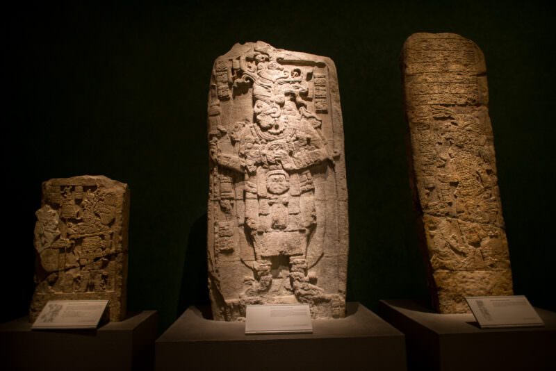 Three carved limestone Maya stelae, central figure depicting richly costumed ruler flanked by glyphic columns