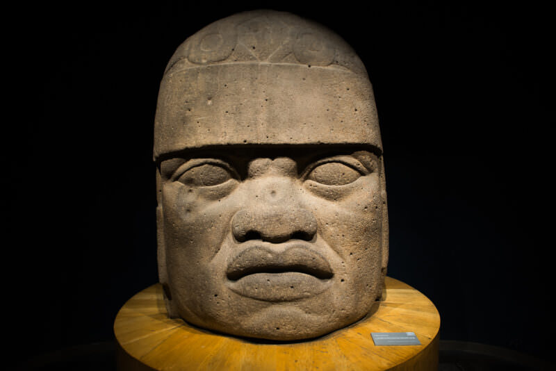 Olmec colossal basalt head spotlit in Gulf of Mexico hall, distinctive facial features and headgear emblems visible