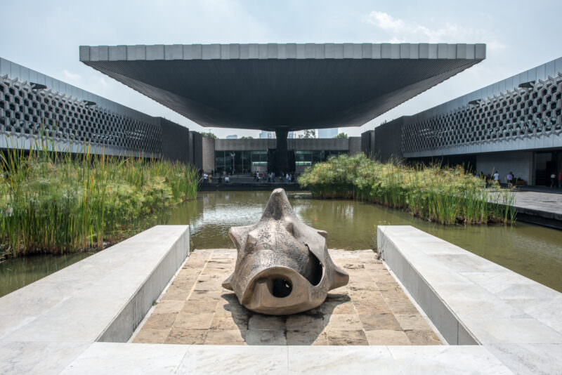 El paraguas cantilevered canopy rising over courtyard from Paseo de la Reforma approach, bronze sculpture in foreground, papyrus-lined reflecting pools