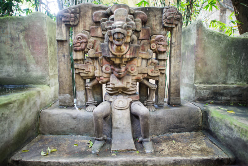 Stone deity sculpture labeled Dios de la Muerte outdoors, flanked by skull-like faces and surrounded by tropical bamboo